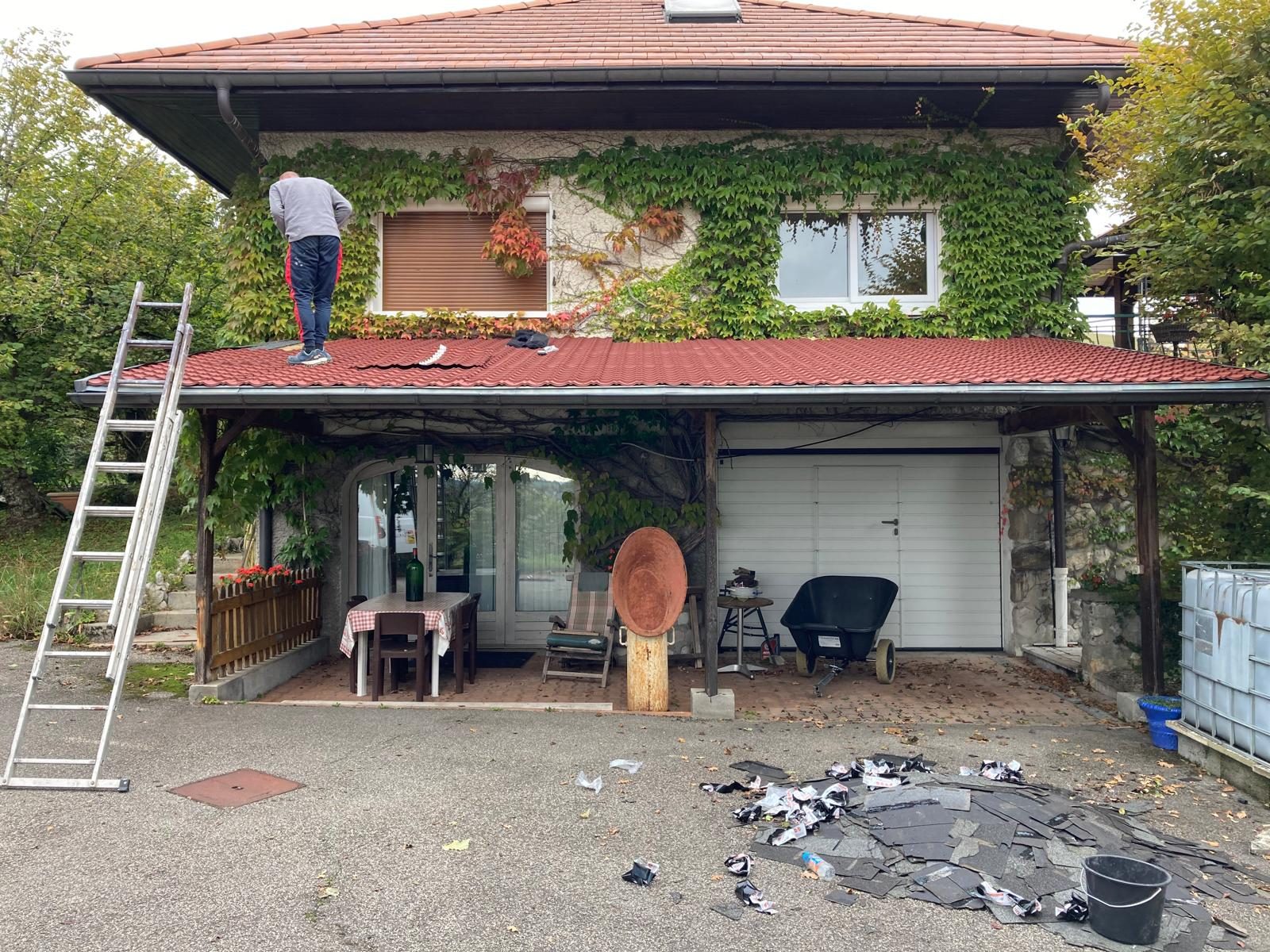 Un couvreur Hoffmann sur la toiture d'un auvent, devant une maison couverte de vigne vierge